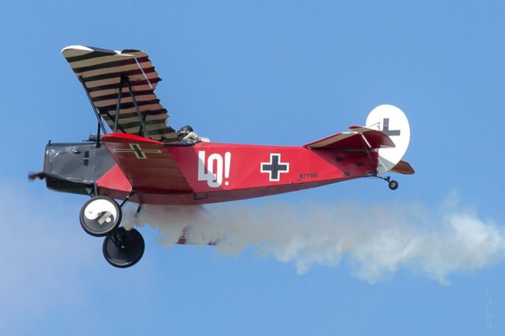 Red Fokker D.VII replica with a striped top wing and LO! painted on the side, trailing white smoke across a blue sky