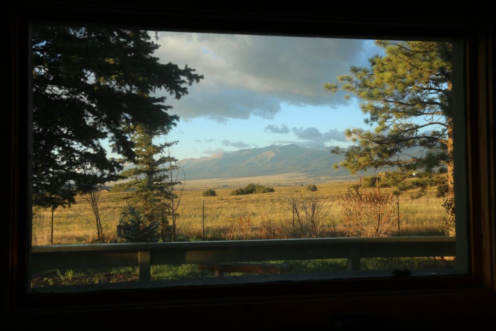 View through a dark window frame of golden grassland, pines and distant mountains under clouds