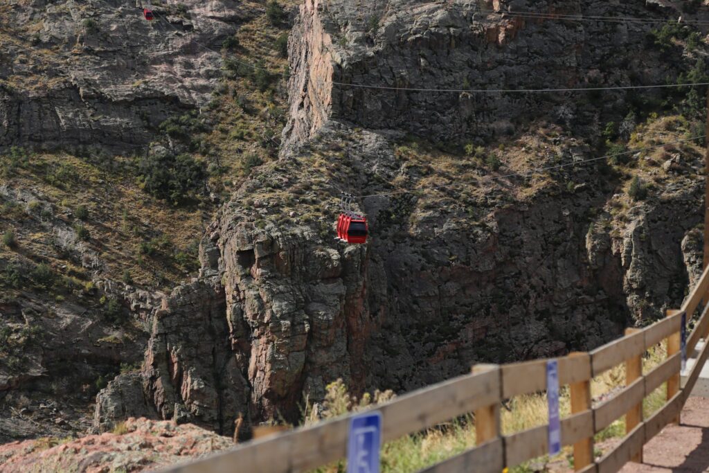 Red gondola cars on cables crossing a deep rocky gorge, seen past a wooden fence