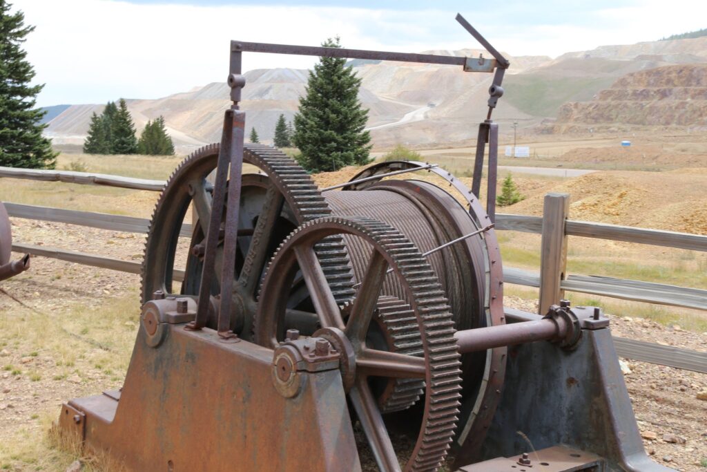 Rusty old mine hoist with large gears and a cable drum with mine tailings in the background
