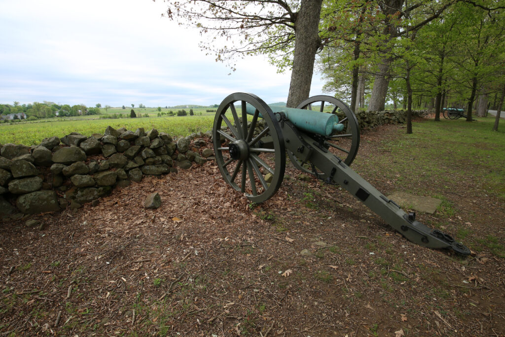 Green bronze cannon behind a low stone wall in the woods, looking out over an open field