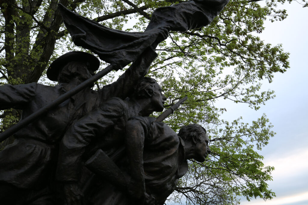 Close-up of bronze Confederate soldiers charging forward under a flag, against spring leaves and sky, North Carolina State Memorial