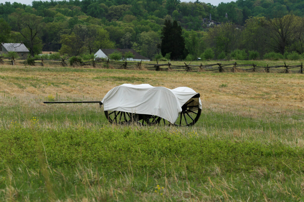 Wagon with a white canvas cover sitting in a tan hayfield, with a split-rail fence and farm buildings behind
