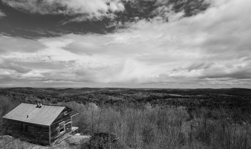 Weathered wooden cabin on a hillside above miles of forested hills, in black and white