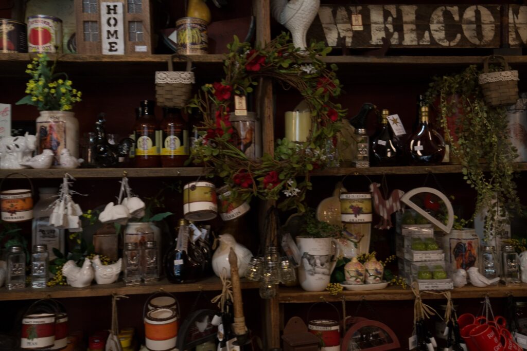 Gift shop shelves packed with country decor: white ceramic hens, jars, a red flower wreath, and HOME and WELCOME signs