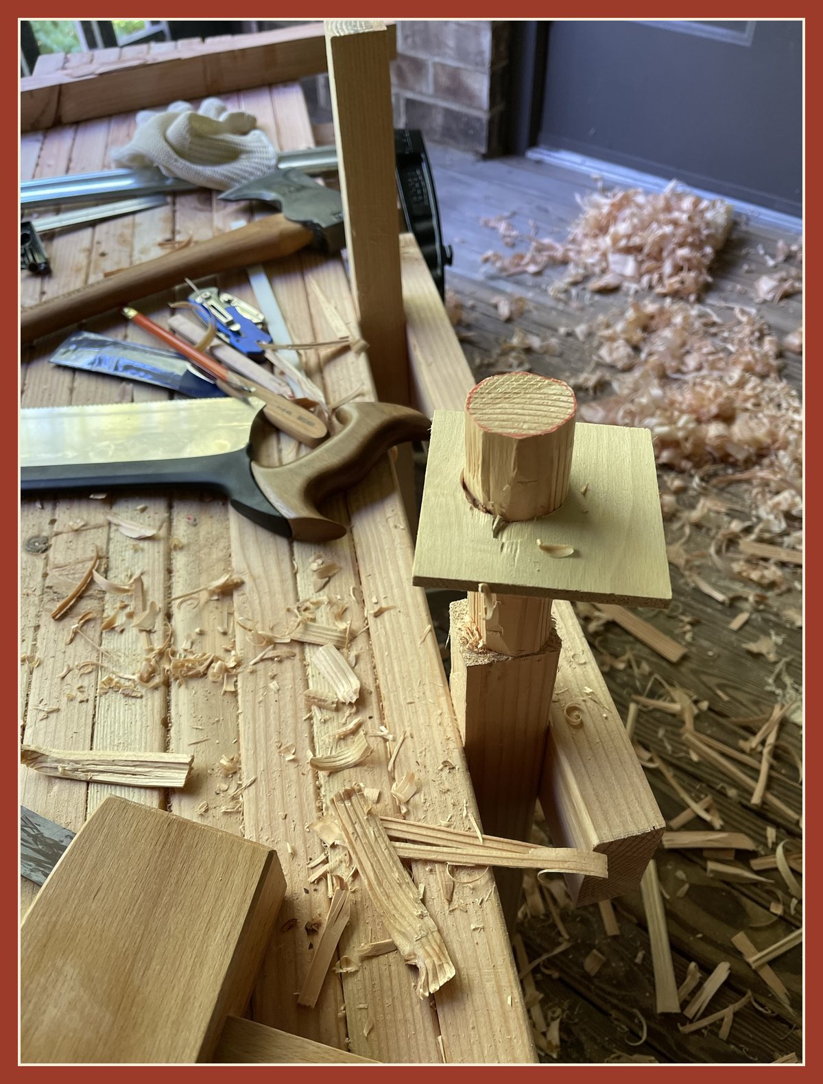 A leg clamped upright with its round tenon poking through a scrap board that has a test hole drilled in it, tools and shavings all around.