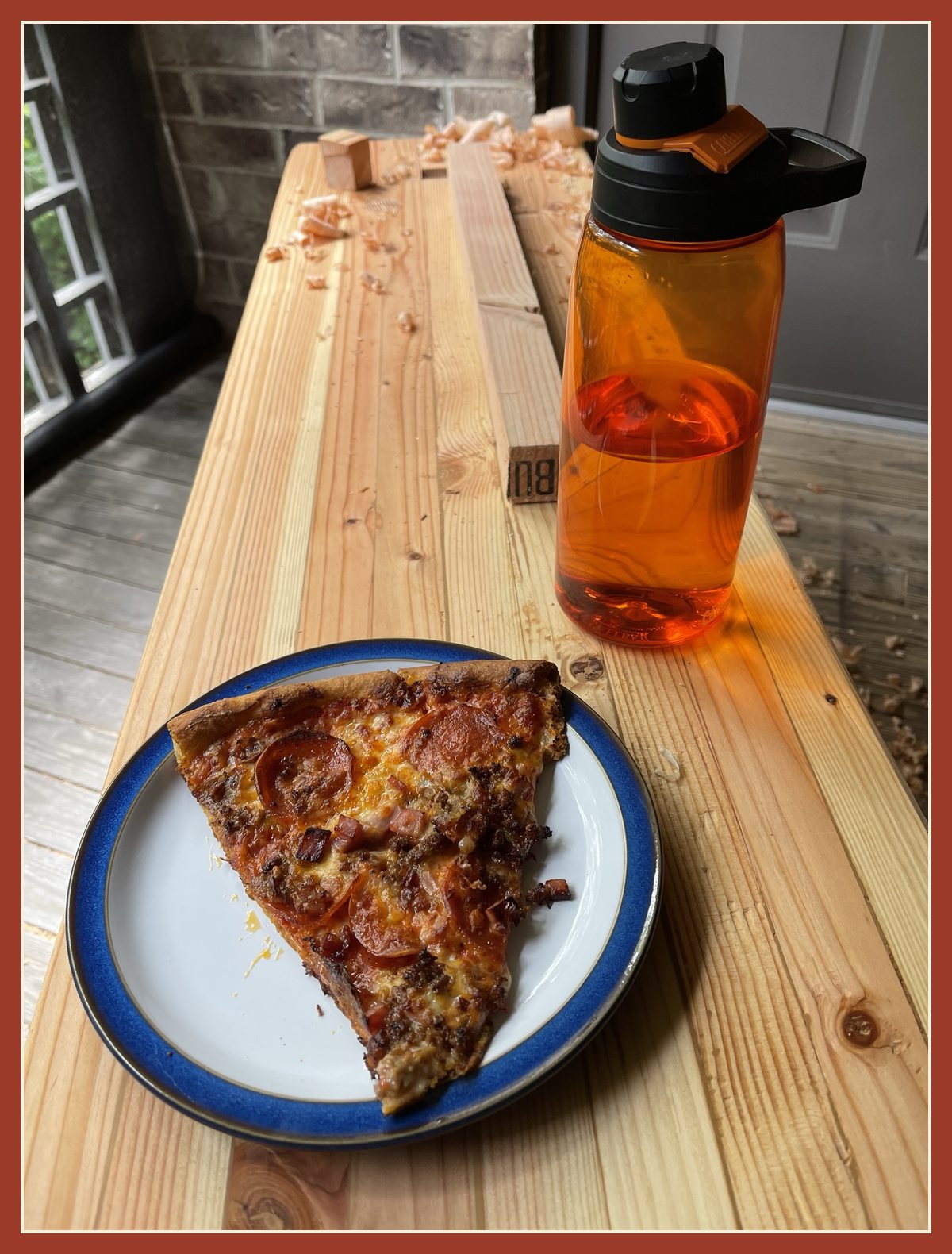 A slice of pepperoni pizza on a blue-rimmed plate and an orange water bottle on the bench top.